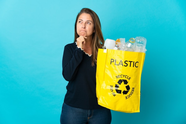Brasilianische Frau mittleren Alters, die eine Tasche voller Plastikflaschen hält, um auf der blauen Wand zu recyceln, die Zweifel und Denken hat