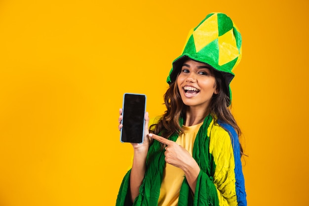 Brasilianischer Fan. Porträt, brasilianischer Fan, der ihr Handy zeigt, gekleidet als Fußballfan oder Fußballspiel auf gelbem Hintergrund. Farben Brasiliens.WM