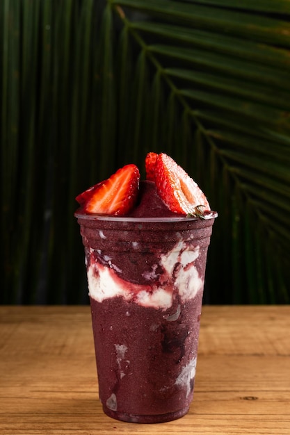 Brasilianischer Frozen Açai Berry Ice Cream Smoothie im Plastikbecher mit Erdbeeren, Bananen und Kondensmilch. Auf Sommer verlässt Hintergrund. Frontansicht für Speisekarte und Social Media