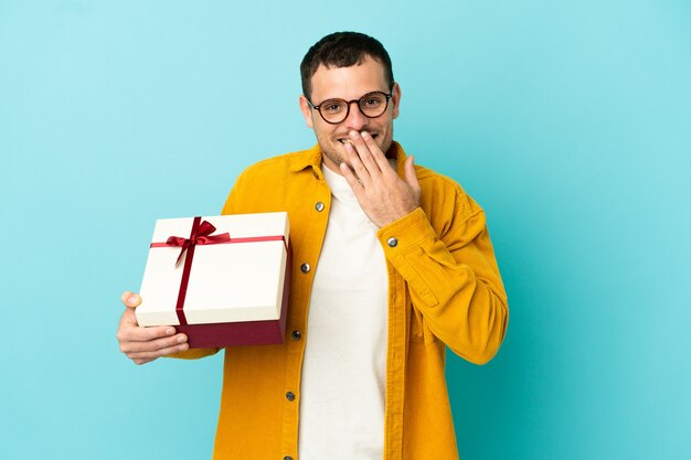 Brasilianischer Mann, der ein Geschenk über isoliertem blauem Hintergrund hält, glücklich und lächelnd, den Mund mit der Hand bedeckend