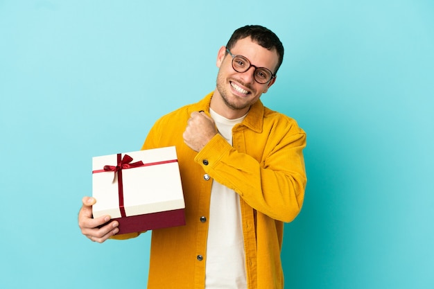 Brasilianischer Mann, der ein Geschenk über isoliertem blauem Hintergrund hält und einen Sieg feiert