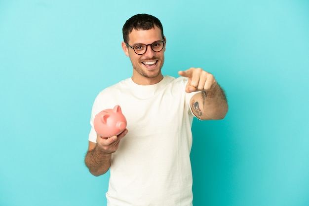 Brasilianischer Mann, der ein Sparschwein über isoliertem blauem Hintergrund hält, überrascht und zeigt nach vorne