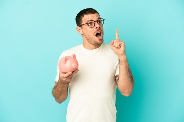 Brasilianischer Mann, der ein Sparschwein über isoliertem blauem Hintergrund hält und eine Idee denkt, die mit dem Finger nach oben zeigt