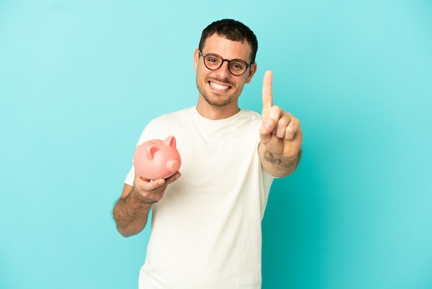 Brasilianischer Mann, der ein Sparschwein über isoliertem blauem Hintergrund hält und einen Finger zeigt und hebt