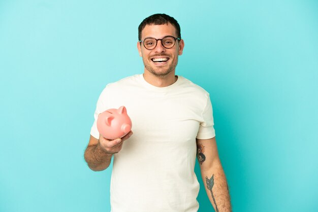 Brasilianischer Mann, der ein Sparschwein über isoliertem blauem Hintergrund mit überraschendem Gesichtsausdruck hält