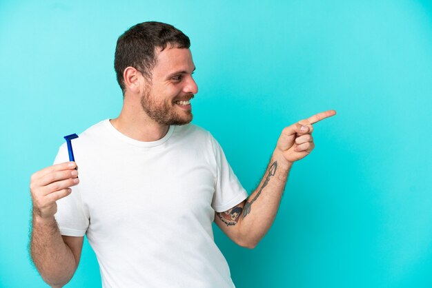 Brasilianischer Mann, der seinen Bart isoliert auf blauem Hintergrund rasiert, mit dem Finger zur Seite zeigt und ein Produkt präsentiert