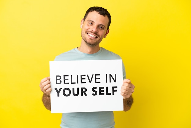 Brasilianischer Mann über isoliertem lila Hintergrund, der ein Plakat mit dem Text Believe In Your Self mit glücklichem Ausdruck hält