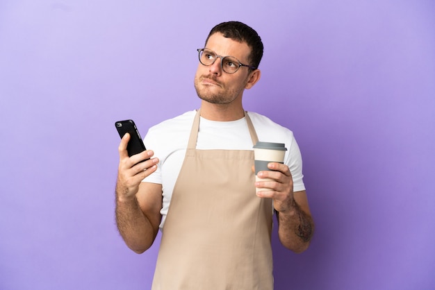 Brasilianischer Restaurantkellner über isoliertem lila Hintergrund, der Kaffee zum Mitnehmen und ein Handy hält, während er etwas denkt?