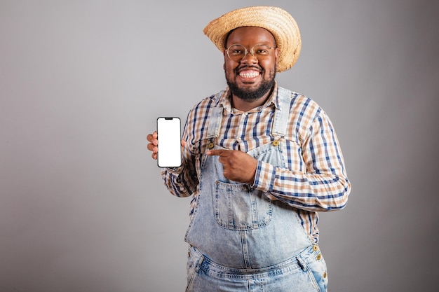 Brasilianischer schwarzer Mann in ländlicher Kleidung von festa junina festa de sao joao arraia hält ein Smartphone mit leeren Bildschirm-Apps auf dem Handy