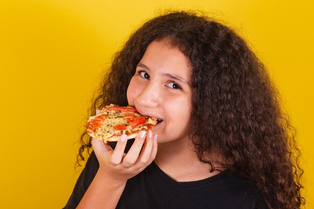 Brasilianisches lateinamerikanisches Mädchen für Afro-Haare, gelber Hintergrund, das köstliche Mini-Pizza isst und beißt Pizza-Margarita-Pizza-Käse, der Käse ausdehnt