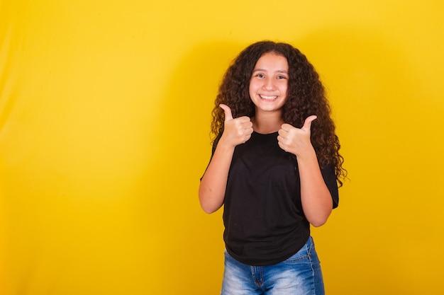 Brasilianisches lateinamerikanisches Mädchen für Afro-Haare, gelber Hintergrund, lächelnde Daumen hoch, ok, positive Bestätigung, Schmuckdaumen