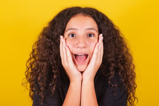 Brasilianisches lateinamerikanisches Mädchen für Afro-Haare, gelber Hintergrund mit der Hand auf dem Gesicht am Kinn, Ausdruck der Überraschung, überraschte, fröhliche, lebhafte, freudige, verängstigte Augen weit