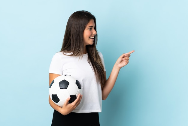 Brasilianisches Mädchen des jungen Fußballspielers lokalisiert auf der blauen Wand, die Finger zur Seite zeigt und ein Produkt präsentiert