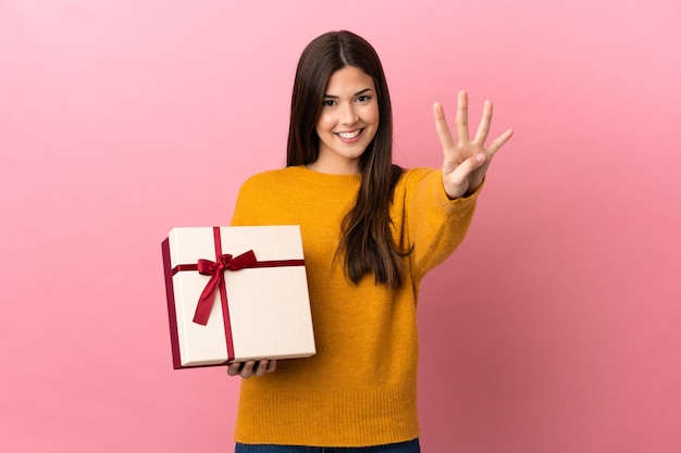 Brasilianisches Mädchen des Teenagers, das ein Geschenk über lokalisiertem rosa Hintergrund glücklich hält und vier mit den Fingern zählt