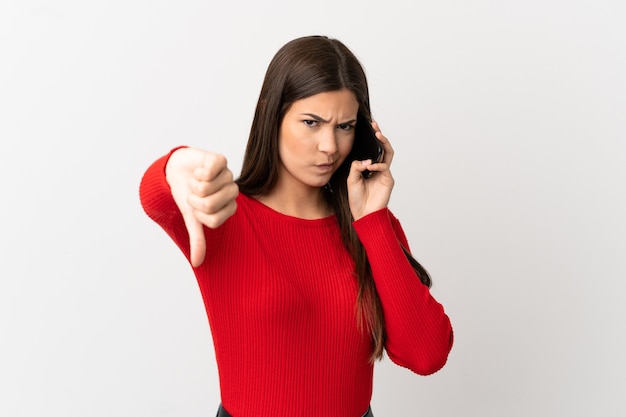 Brasilianisches Mädchen des Teenagers, das Handy über lokalisiertem weißem Hintergrund benutzt, der Daumen nach unten mit negativem Ausdruck zeigt