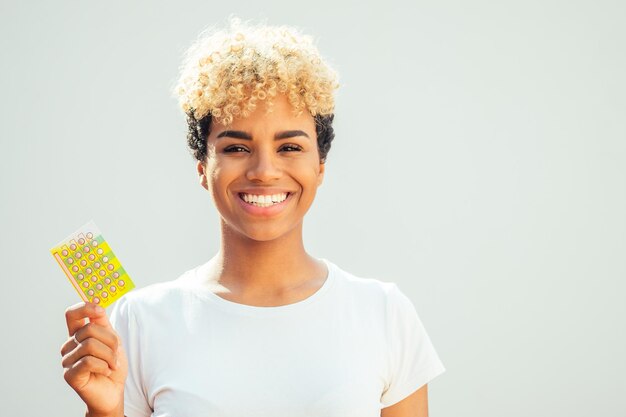 Brasilianisches Mädchen mit niedlichen afroblonden Locken, die Verhütungspillen auf weißem Studiohintergrund zeigen