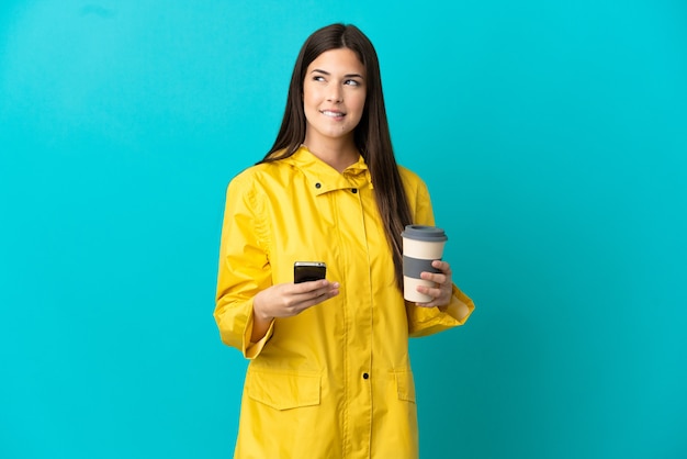 Brasilianisches Teenager-Mädchen, das einen regendichten Mantel über isoliertem blauem Hintergrund trägt, der Kaffee zum Mitnehmen und ein Handy hält, während er etwas denkt