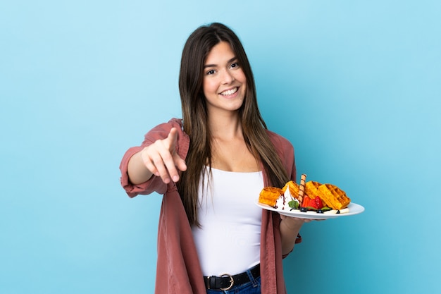 Brasilianisches Teenager-Mädchen, das Waffeln über lokalisiertem blauem Hintergrund hält, zeigt Finger auf Sie mit einem selbstbewussten Ausdruck