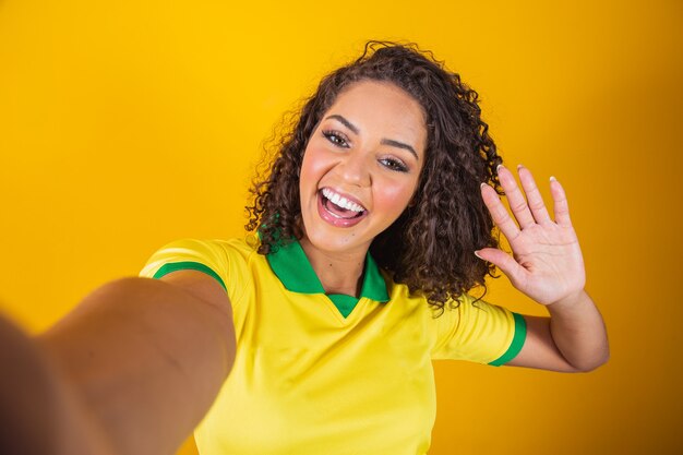 Brasilien-Anhänger. Brasilianischer Frauenfan, der auf Fußball feiert, Fußballspiel auf gelbem Hintergrund. Brasilien farben. Selfie-Smartphone.