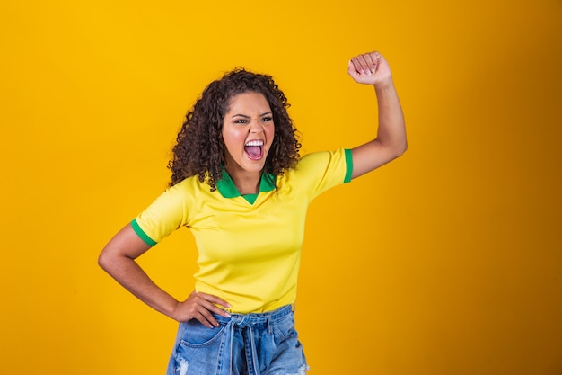 Brasilien-Anhänger. Brasilianischer Frauenfan mit lockigem Haar, der auf Fußball feiert, Fußballspiel auf gelbem Hintergrund. Brasilien farben.