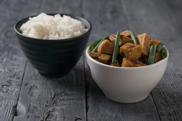 Braten sie tofu in einer weißen schüssel und reis in einer schwarzen schüssel auf einem holztisch. vegetarisches asiatisches gericht.