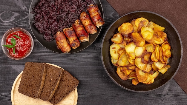 Bratkartoffel im Topf, Roggenschwarzbrotstücke auf einer hölzernen Servierplatte, hausgemachte Würstchen im Speck und gedünsteter Kohl auf schwarzem rustikalem Hintergrund.