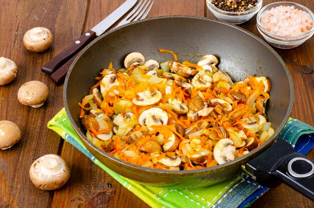 Bratpfanne mit Champignons, Zwiebeln und Karotten mit gebratenen Pilzen auf Holztisch.