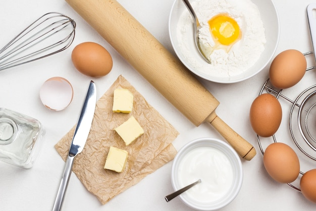 Braune Eier auf Metallständer. Butter und Messer auf Papier. Eigelb mit Mehl und Löffel in eine Schüssel geben. Milch in Schüssel und Nudelholz auf dem Tisch. Weißer Hintergrund. Flach liegen