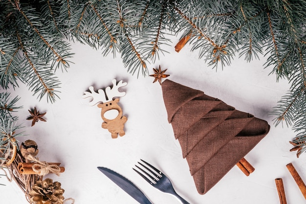 Braune Leinenserviette gefaltet in Form von Weihnachtsbaum und Messer, Gabel auf weißer Oberfläche mit Fichtenzweigen. Ansicht von oben.