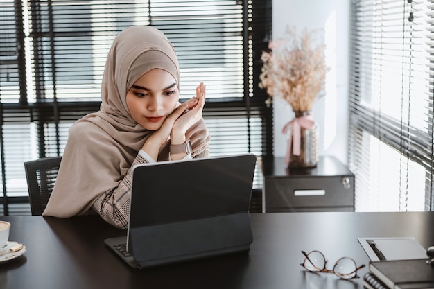 Brauner Hijab der jungen asiatischen muslimischen Geschäftsfrau, der mit Laptop-Computer am modernen Büro sitzt und arbeitet.