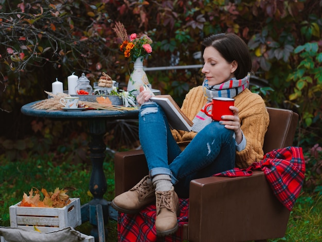 Brauner Mantel der jungen Frau sitzt Stuhl-Tisch-Lesebuch mit Plaid, das über ihrem Kopf unter freiem Himmel gegen herbstrotes Laub geworfen wird