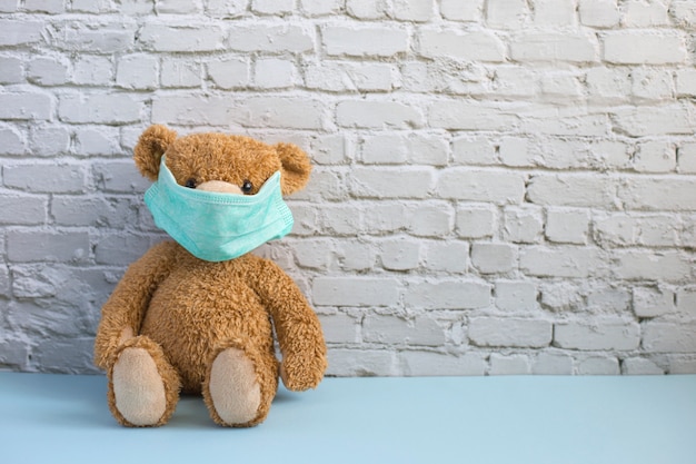 Brauner Teddybär in grüner medizinischer Maske sitzt allein nahe weißer Backsteinmauer. Isolation und canantine Konzept