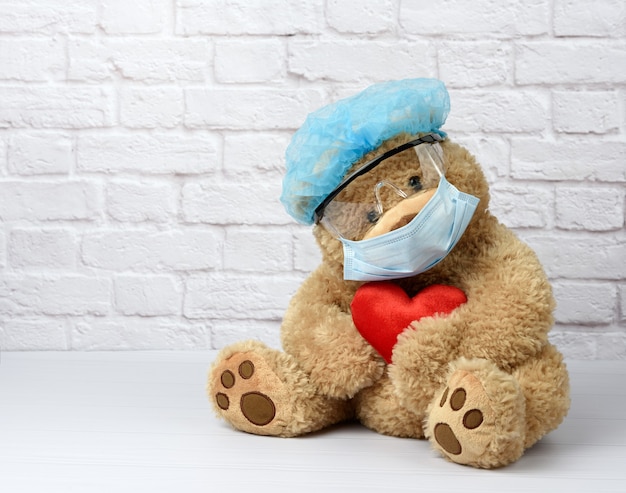 Brauner Teddybär sitzt in einer schützenden Plastikbrille, einer medizinischen Einwegmaske und einer blauen Kappe