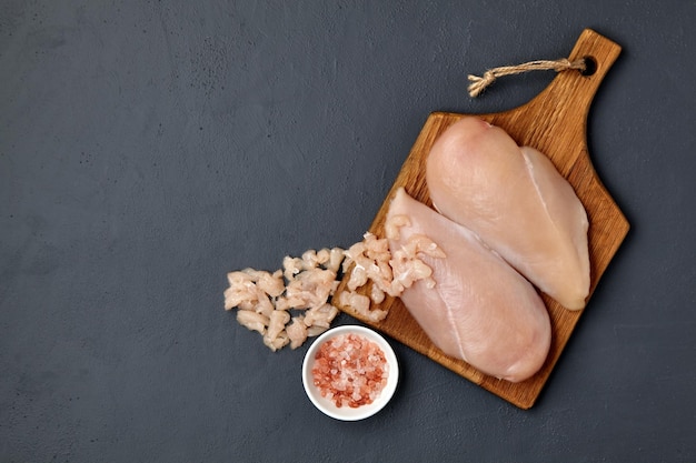 Braunes Holzschneidebrett mit gehacktem Hähnchenbrustfilet und Schüssel mit rosafarbenem Salz auf einem dunklen Betontisch Draufsicht mit Kopierbereich Flach liegend