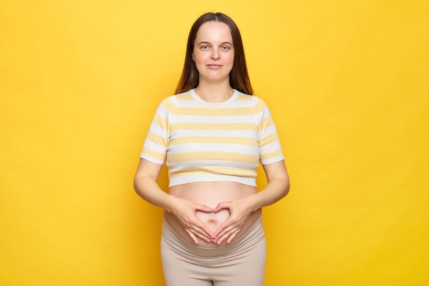 Foto braunhaarige erwachsene schöne kaukasische schwangere frau mit nacktem bauch trägt ein lässiges oberteil, isoliert über gelbem hintergrund, berührt ihren bauch und zeigt ein liebessymbol, das mit den händen gemacht wird