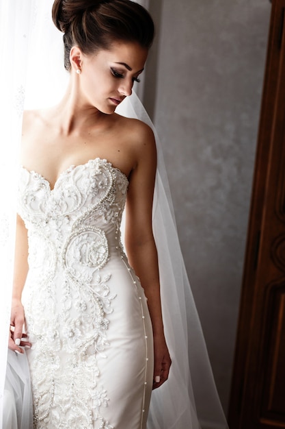 Braut im herrlichen Hochzeitskleid mit crystls und Perlen steht vor hellem Fenster