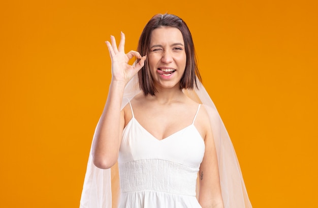 Braut im schönen Hochzeitskleid glücklich und fröhlich, die Zunge herausstreckend, die das okayzeichen zeigt, das über orange Wand steht