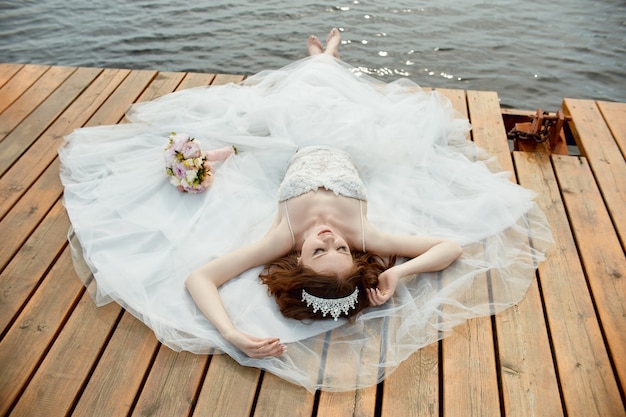 Braut im weißen Kleid steht auf der Brücke und schaut auf den See. Frau im Hochzeitskleid mit Blumenstrauß