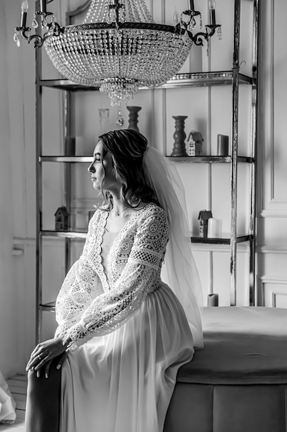 Braut in einem Hochzeitskleid im Boho-Stil und mit einem Schleier posierend in einem gemütlichen Raum sitzend.