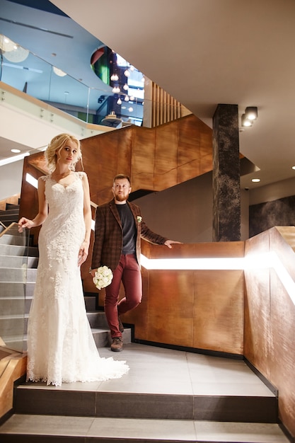 Braut in einem schicken langen Kleid mit einem Zug und dem Bräutigam stehen auf großen Treppen, Paar in Liebesumarmungen auf Treppen küssen sich und schauen sich an