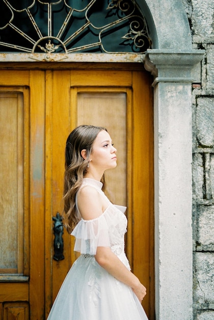 Braut in einem weißen Kleid steht in der Nähe einer Holztür-Seitenansicht