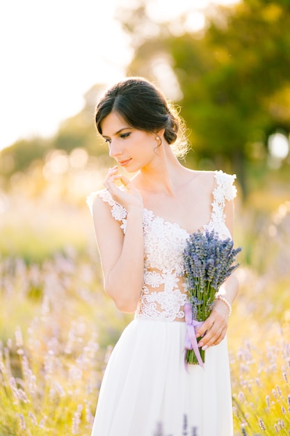 Braut in einem weißen Spitzenkleid steht auf einem Feld und hält in der Hand einen Lavendelstrauß gebunden mit einem