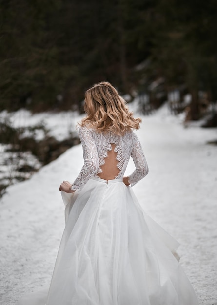 Braut mit blonden Haaren, die im Winter von der Hochzeit in weißem Trachtenkleid mit offenem Rücken davonlaufen