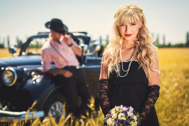 Braut mit Blumenstrauß und Bräutigam nahe altmodischem Auto auf dem Feld