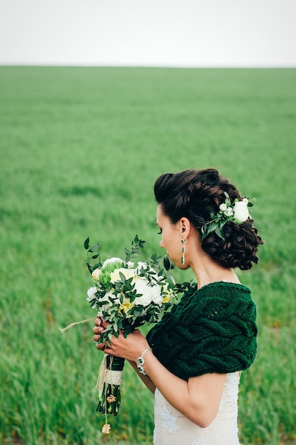 Braut mit einem strauß in einem elfenbeinfarbenen kleid und einem gestrickten schal auf einer grünen wiese