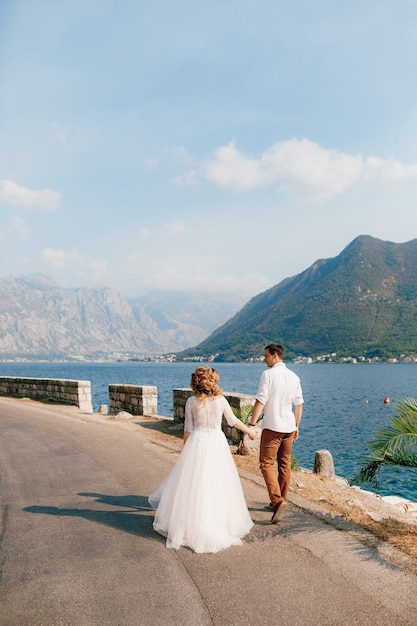 Braut und Bräutigam gehen Hand in Hand die Straße entlang der Küste in der Bucht von Kotor in der Nähe von Perast