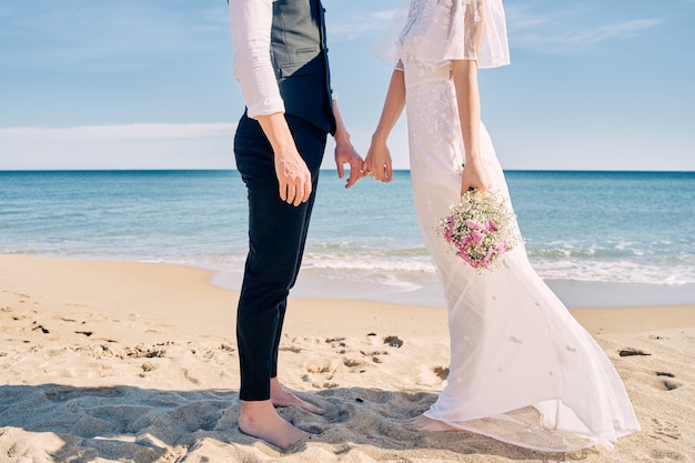 Braut und Bräutigam Händchen haltend am Strand in Brautkleidern