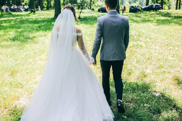 Braut und Bräutigam Händchen haltend im Park Glückliches Paar zu Fuß zusammen Hochzeitstag Foto Liebesgeschichte Schönes langärmliges Kleid Spitzenschleier Stilvoller rustikaler Blumenstrauß