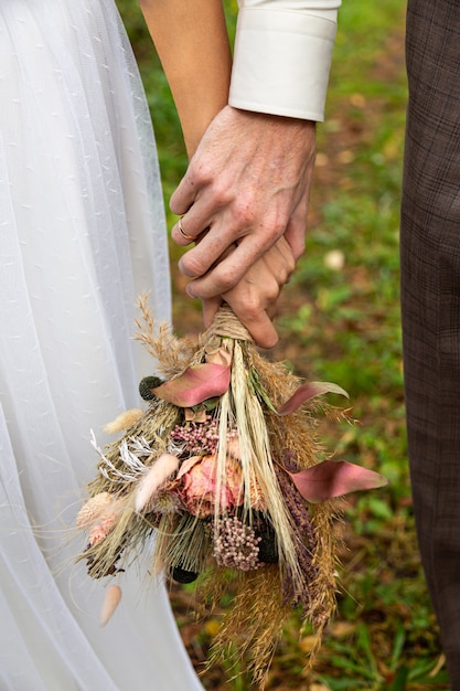 Braut und Bräutigam halten sich an den Händen. In den Händen ein Strauß getrockneter Blumen im Boho-Stil