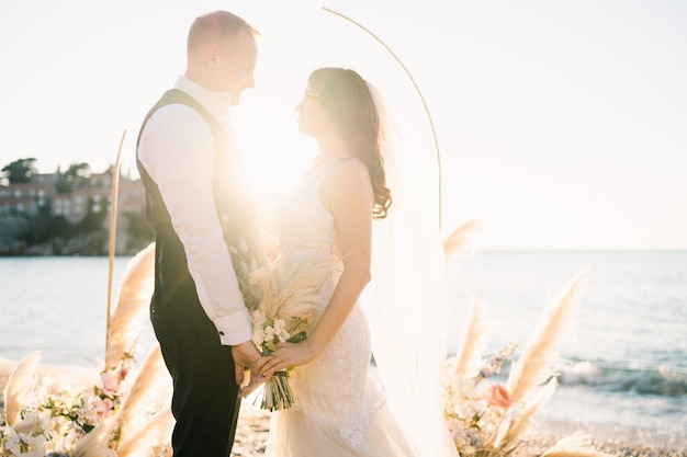 Foto braut und bräutigam halten sich im sonnenlicht in der nähe des hochzeitsbogens am strand an den händen
